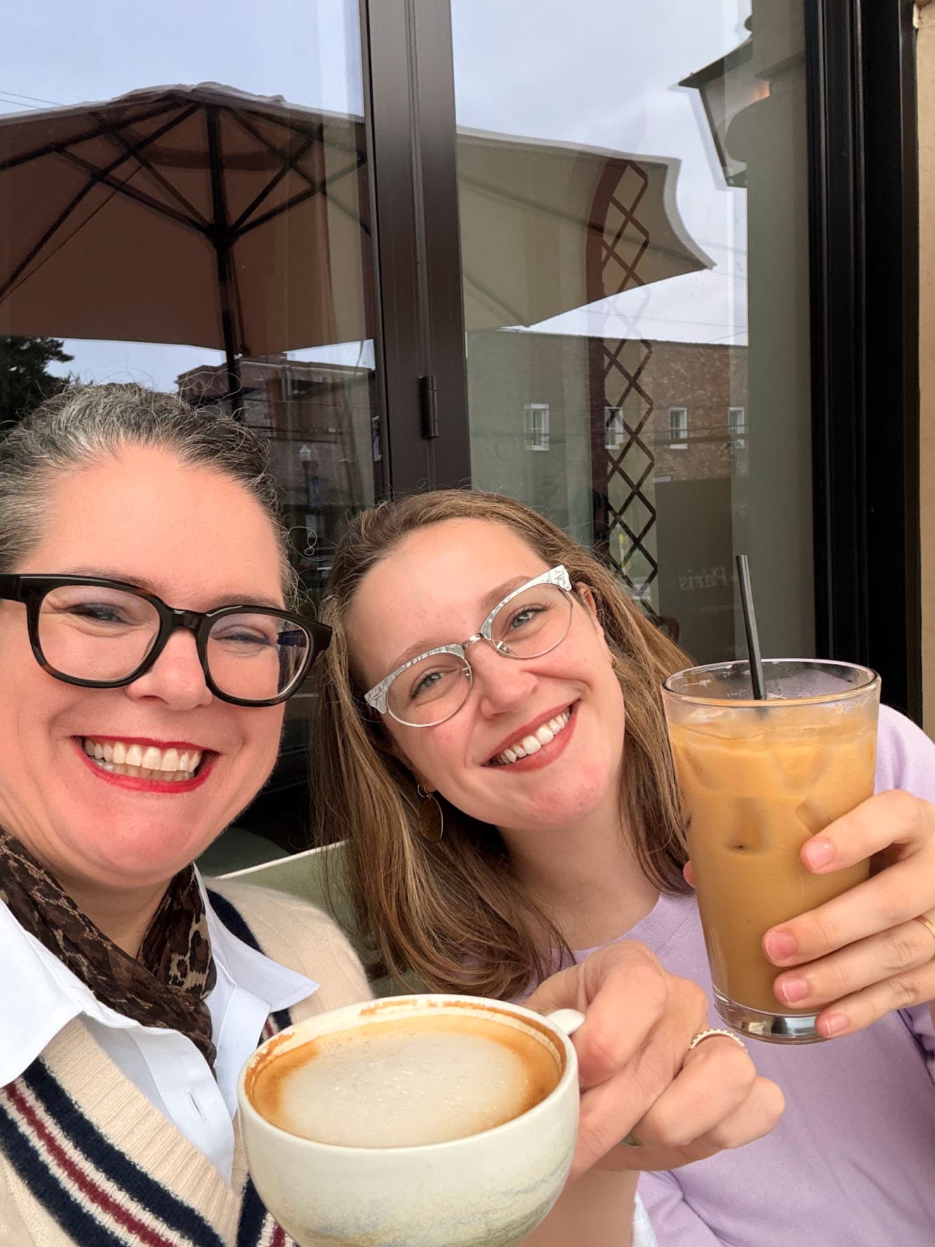 Laura and Kelsey at a café table outside, raising a cappuccino and an iced coffee to the camera.